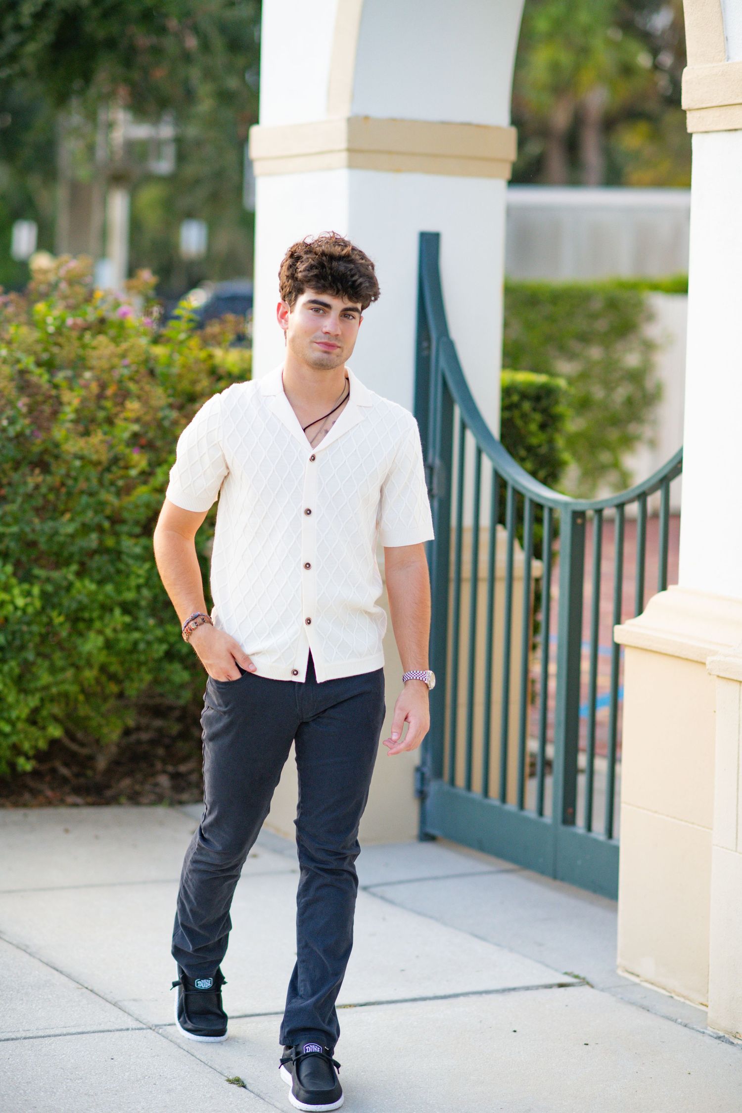 Young man in white button-up shirt and dark jeans posing confidently near decorative gate outdoors.