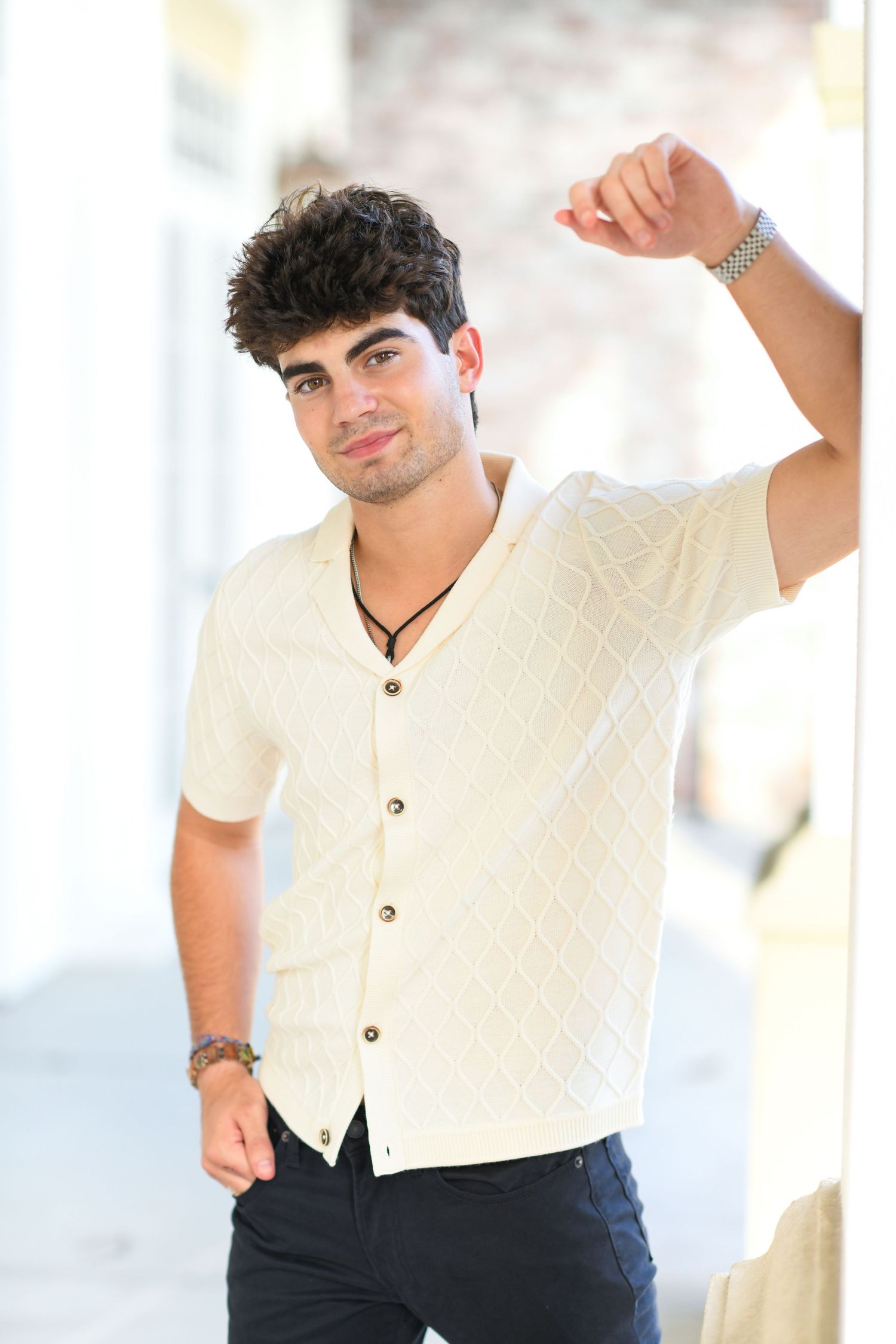 Young man in cream diamond-pattern button shirt leaning against white column.