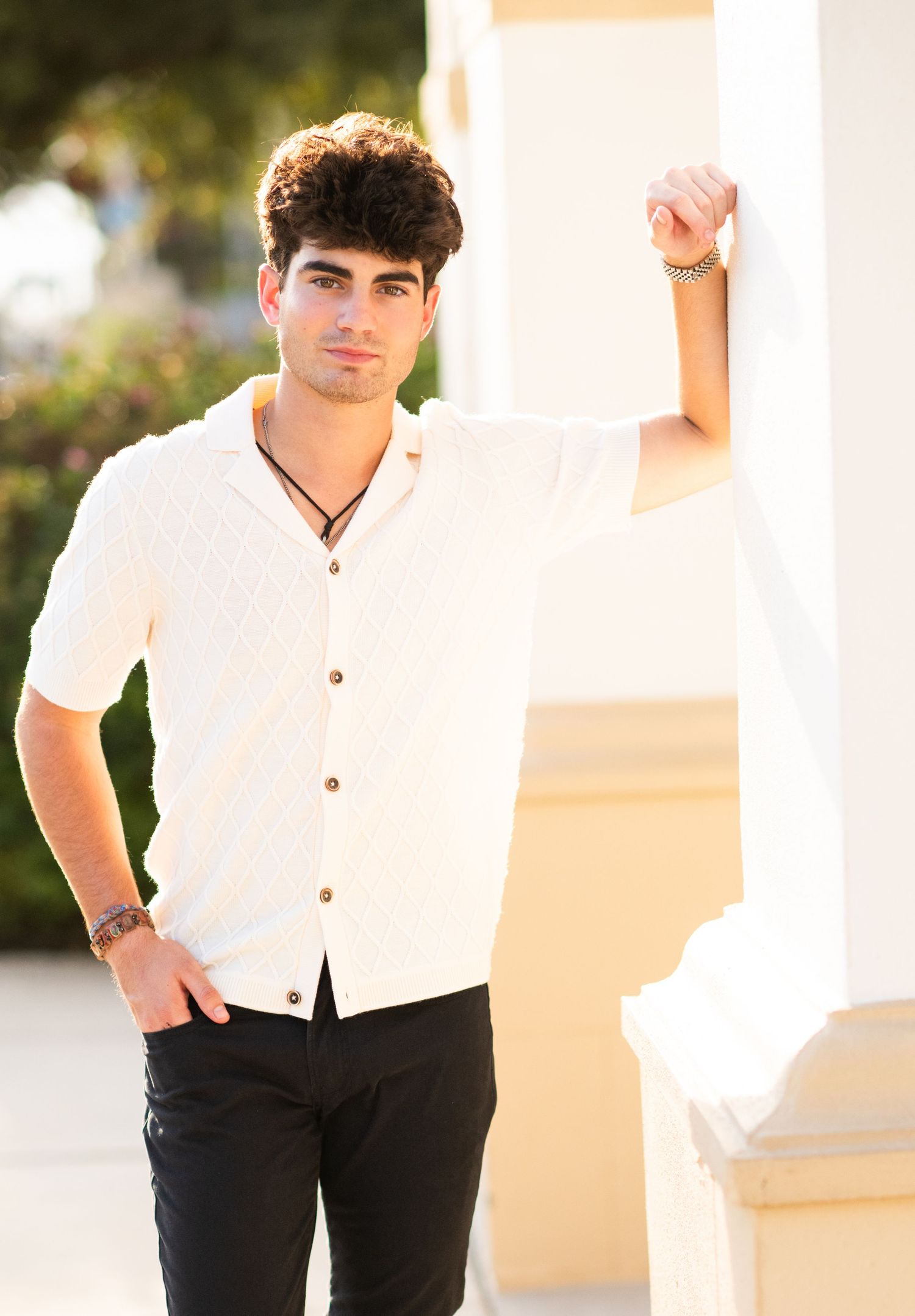 Young man in white button-up shirt and black pants leaning against white column outdoors.