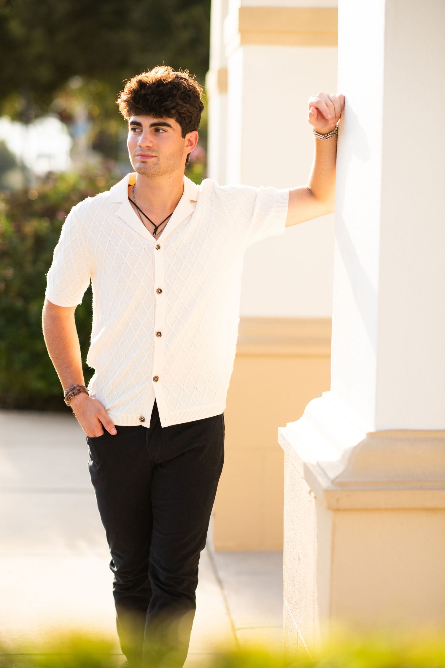 Young man in white short-sleeve shirt and black pants leaning against white column outdoors.