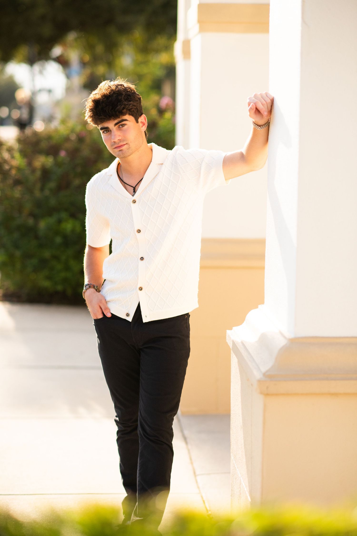 Young man in white shirt and black pants leaning against a pillar outdoors in warm sunlight.