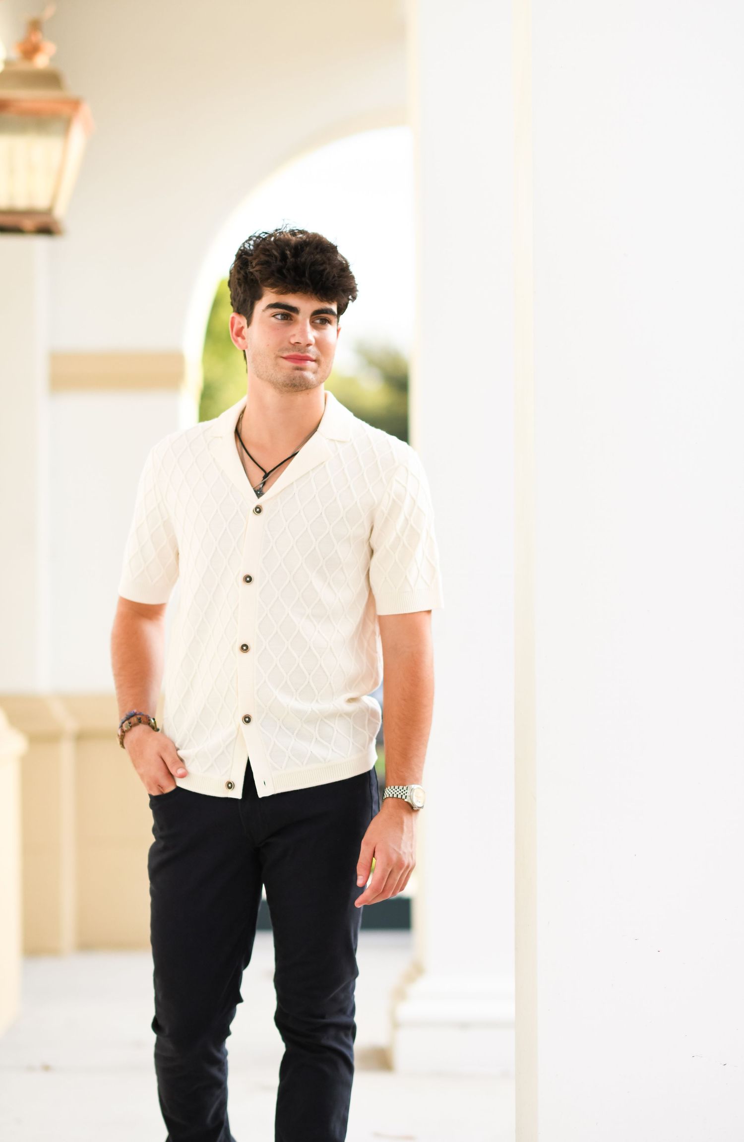 Young man in white button-up shirt and black pants posing between white columns outdoors.