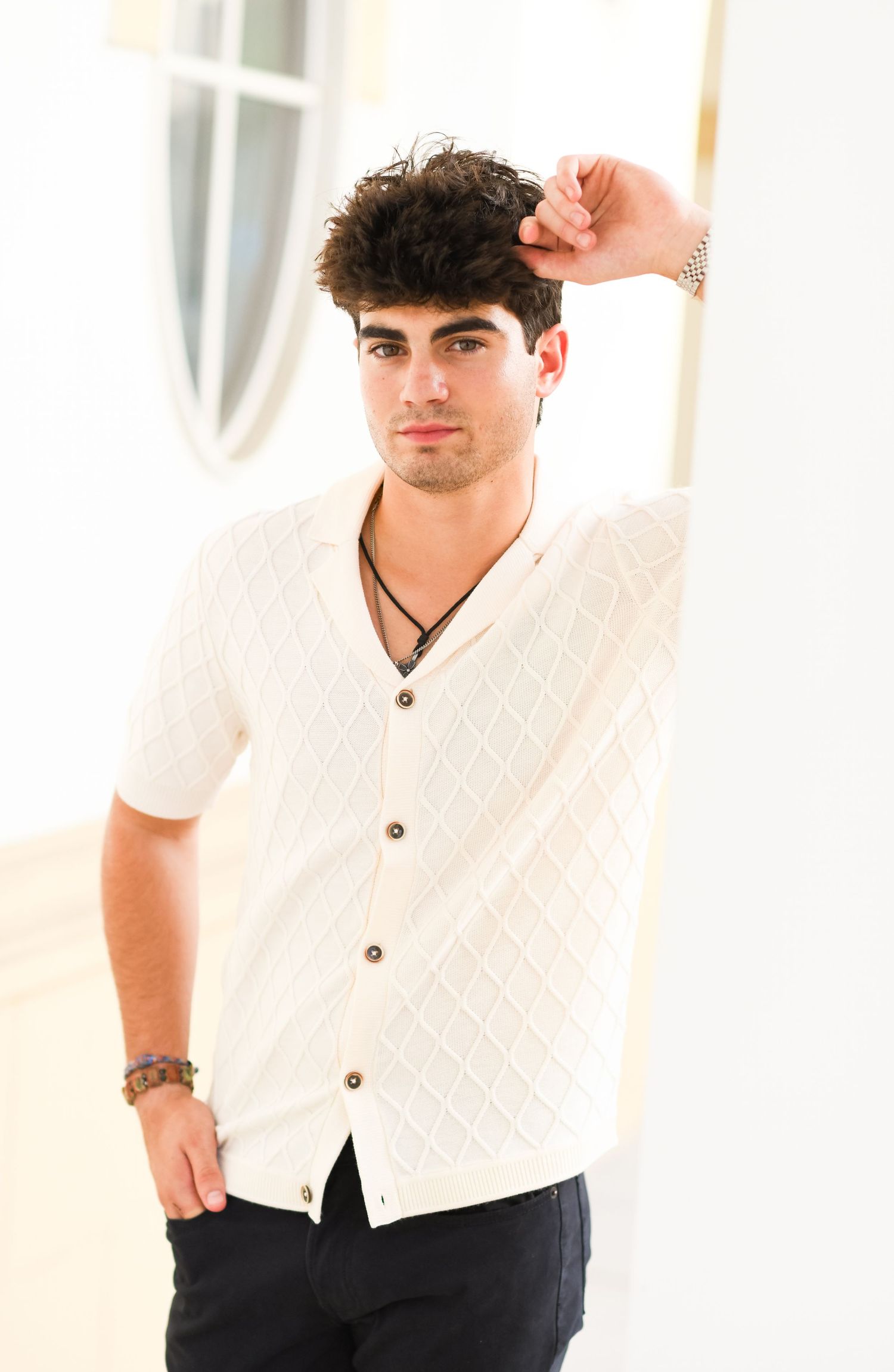Young man in white diamond-knit button-up shirt posing indoors casually.