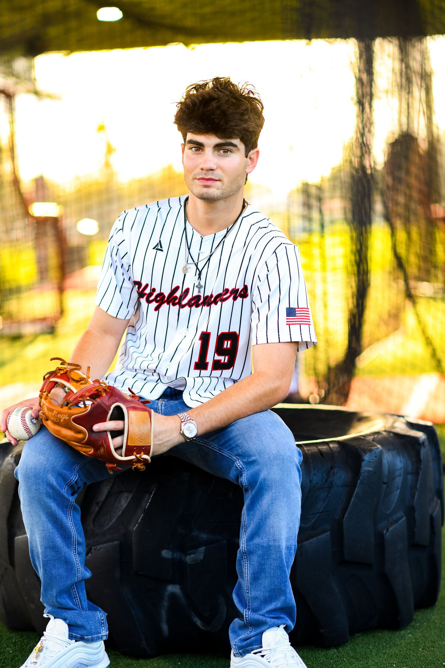 Young baseball player in Highlanders jersey number 19 sitting on tire with glove at field.