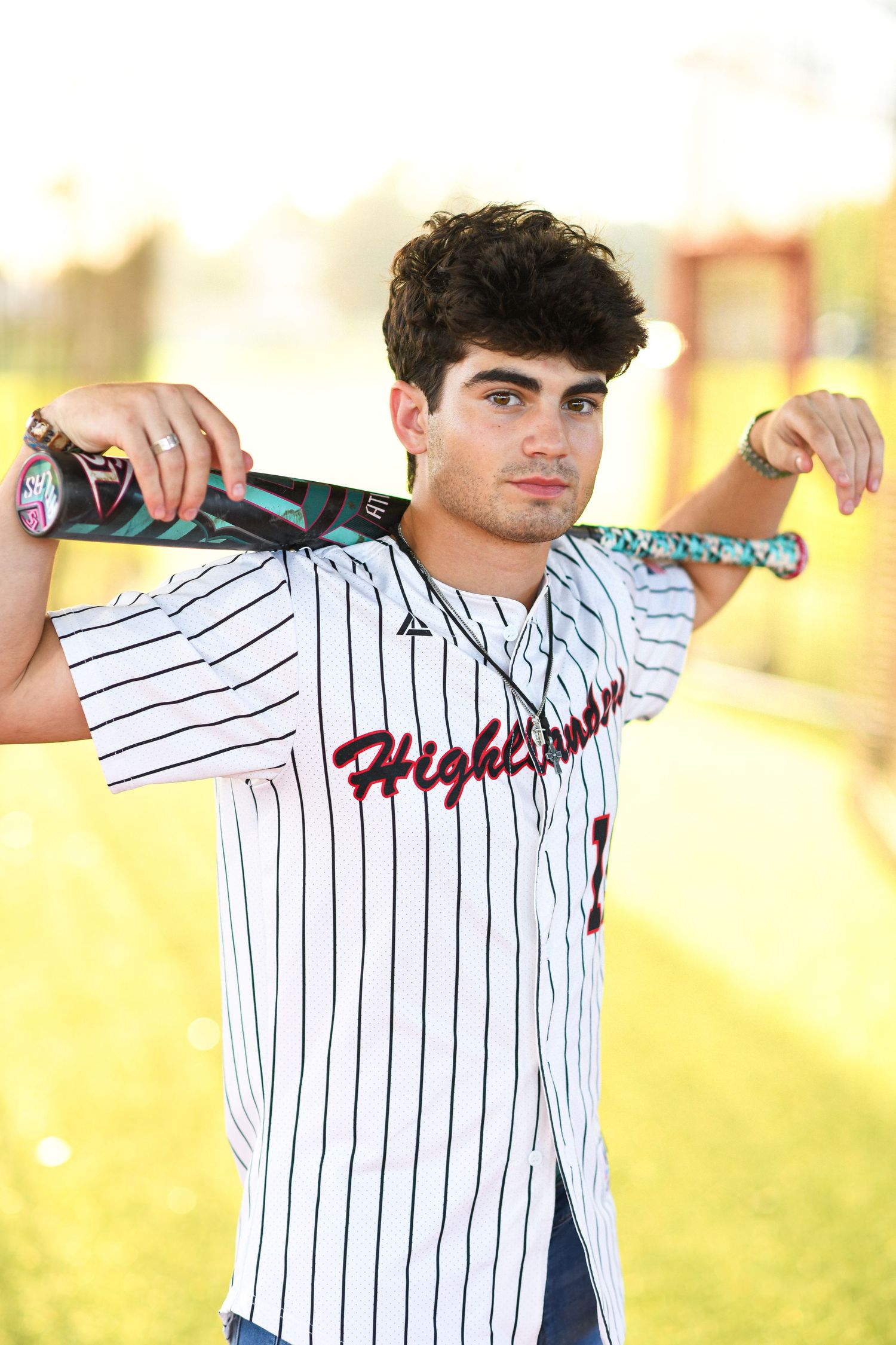 High school baseball player in Highlanders jersey holding bat across shoulders on field.