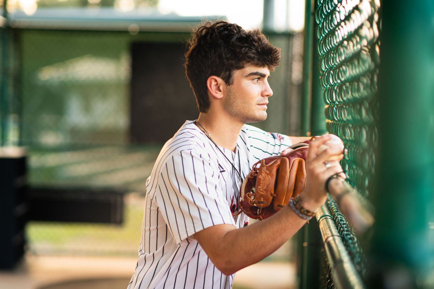 Young baseball player in pinstripe jersey leaning on green chain-link fence at sunset.