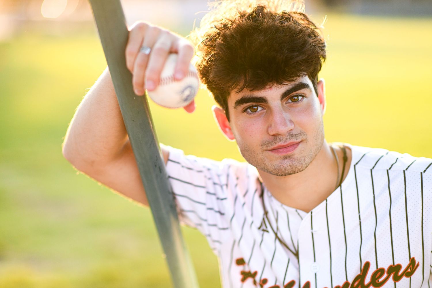 Teen baseball player in pinstripe uniform leaning on pole holding ball in golden hour light.