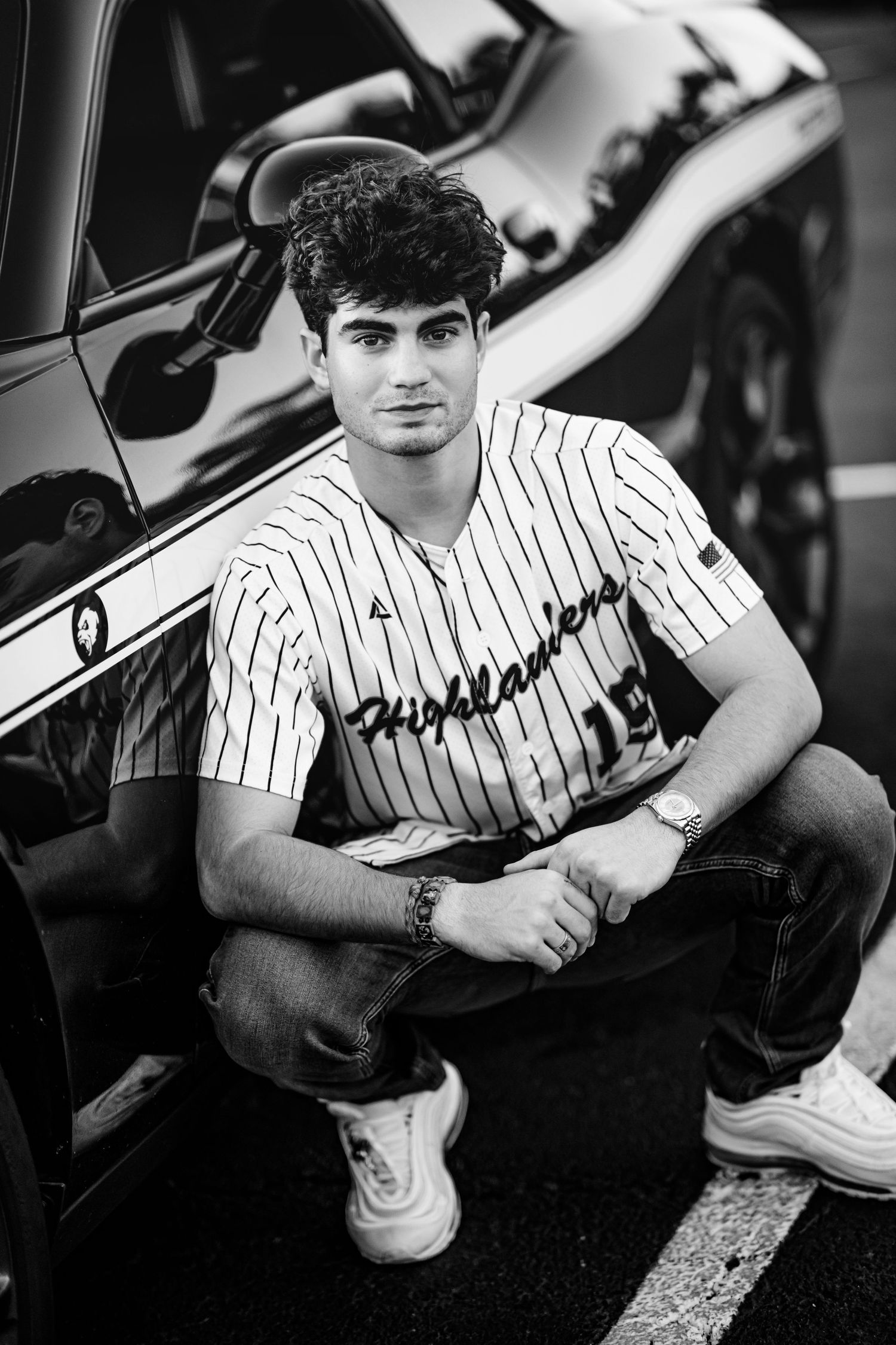 Black and white portrait of young man in Highlanders baseball jersey beside dark car.
