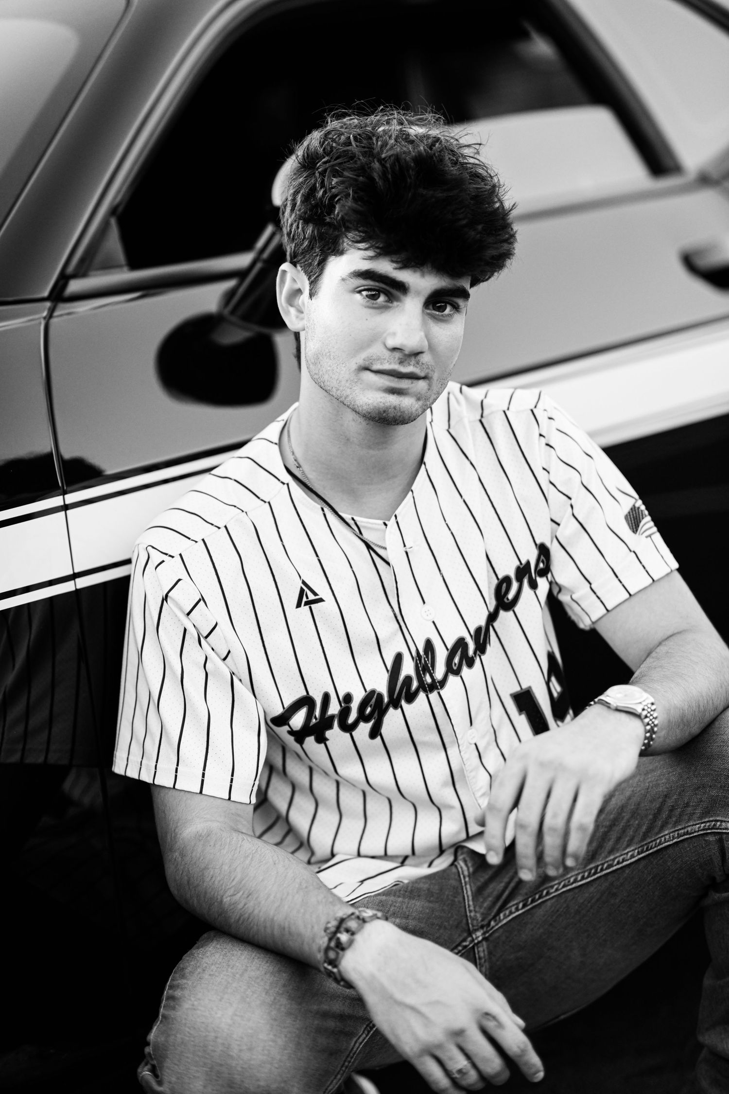 Teen boy in Highlanders baseball jersey sitting against a car in black and white photo.