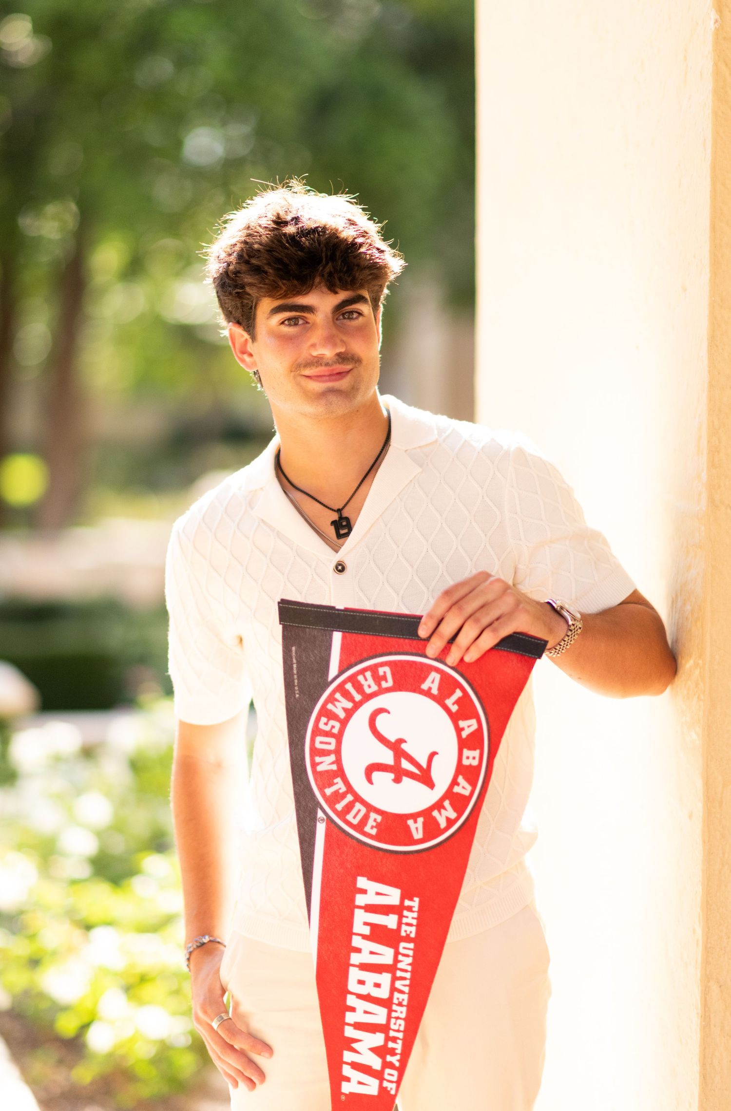 Smiling young man holding University of Alabama pennant while leaning against white column outdoors.