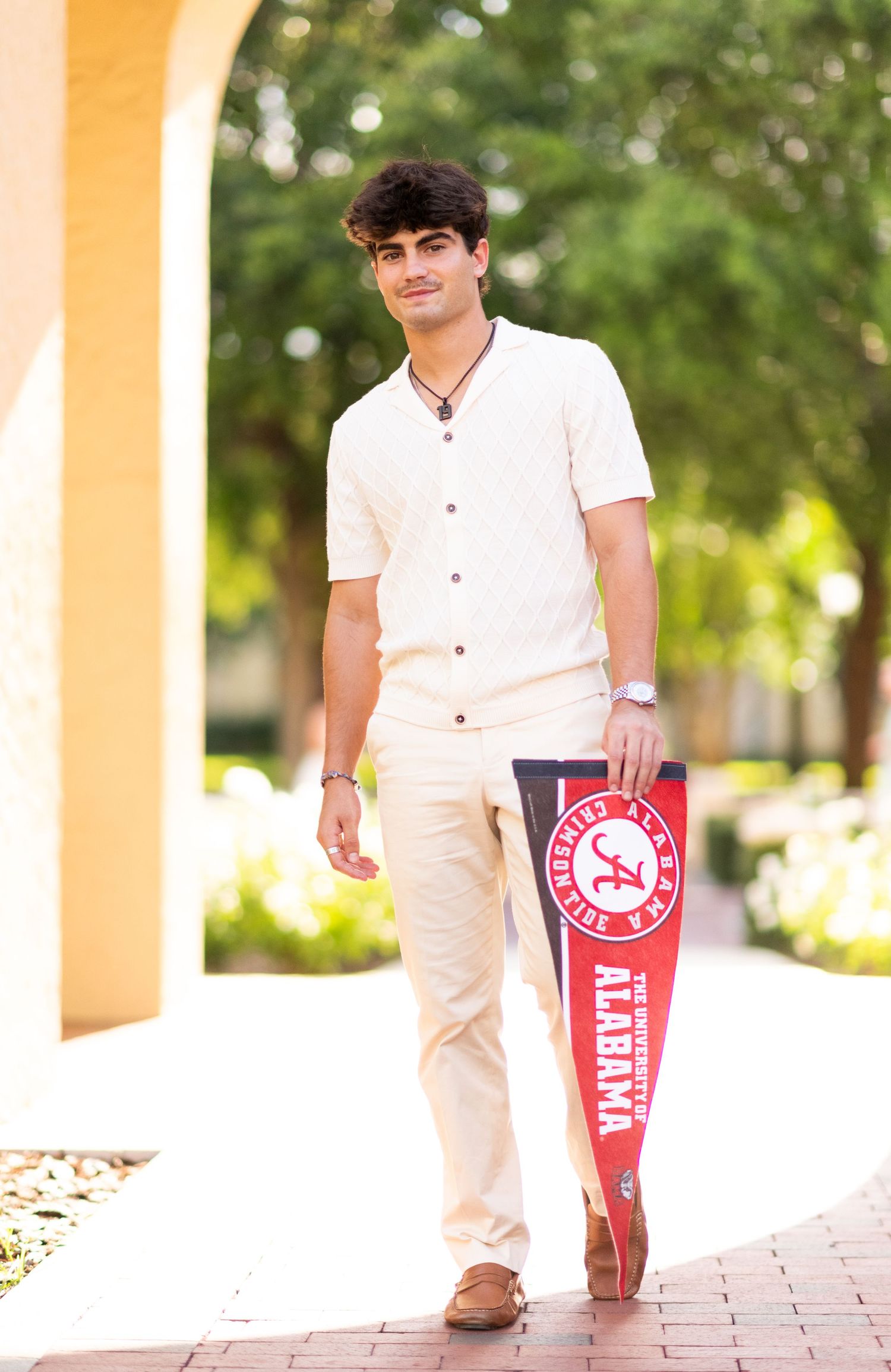 Young man walking outdoors holding a University of Alabama pennant on a brick pathway.