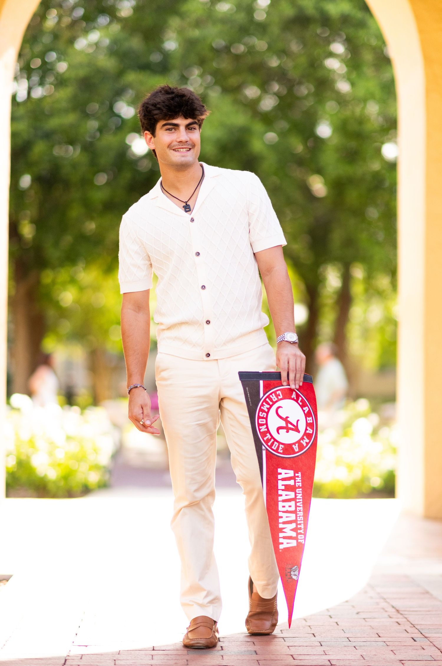 Smiling young man in white shirt holding University of Alabama pennant walking outdoors.
