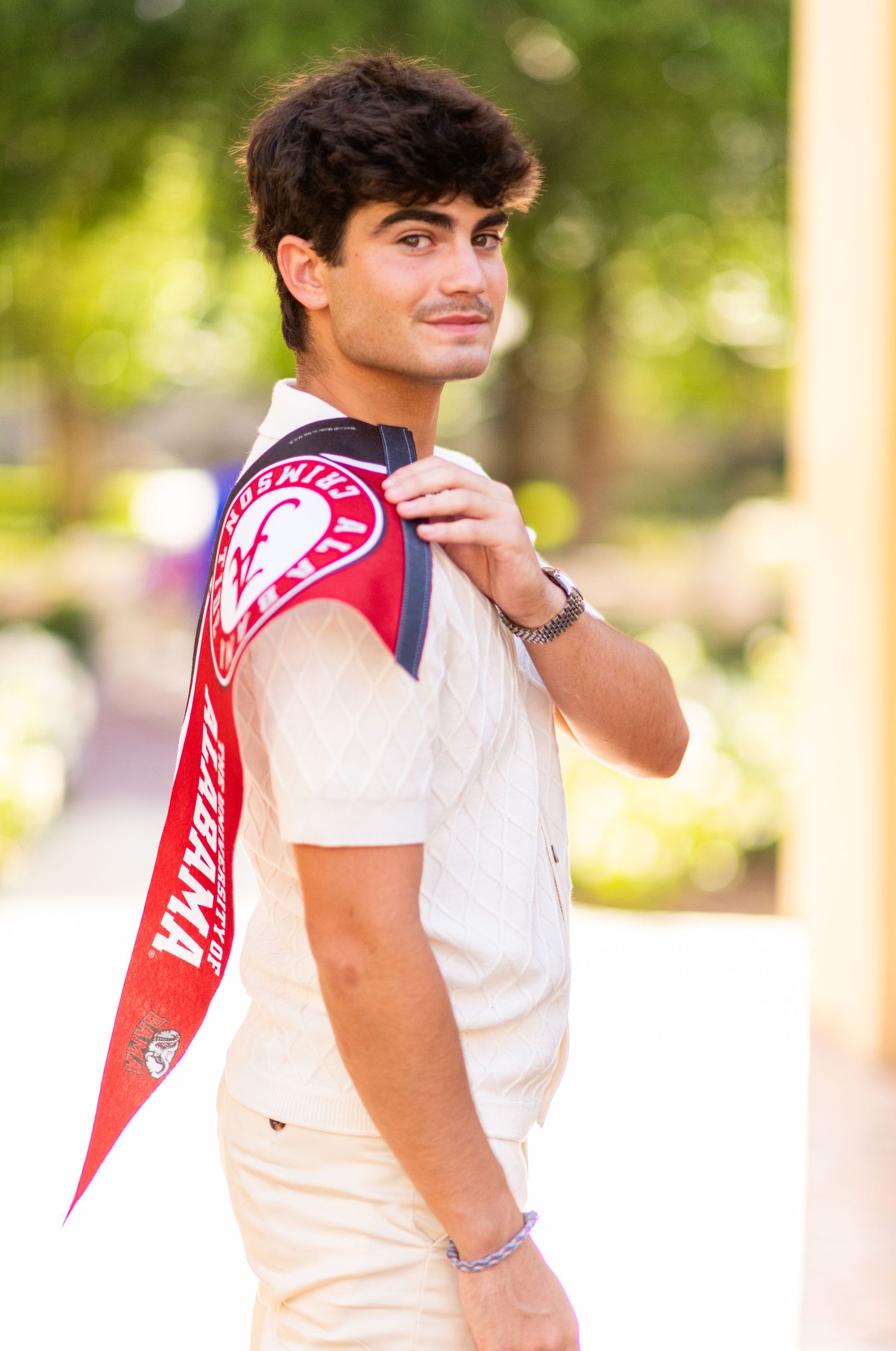 Young man posing outdoors wearing a University of Alabama red sash over a white polo shirt.