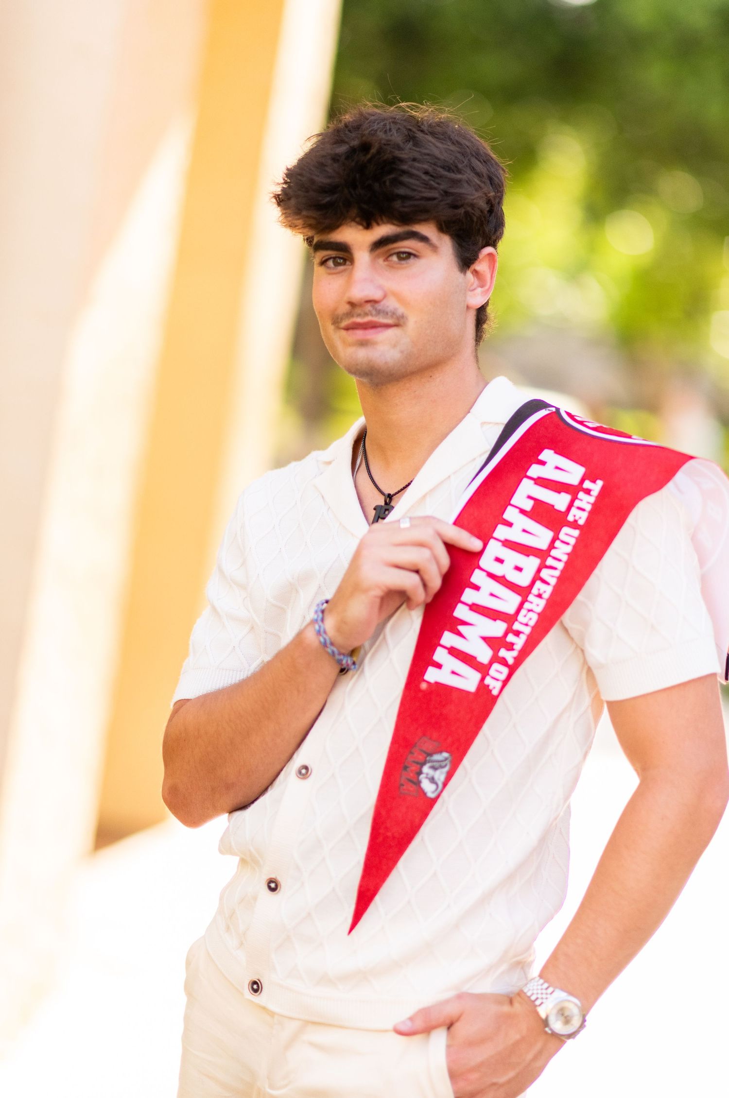 College student holding a University of Alabama red banner sash while posing outdoors.