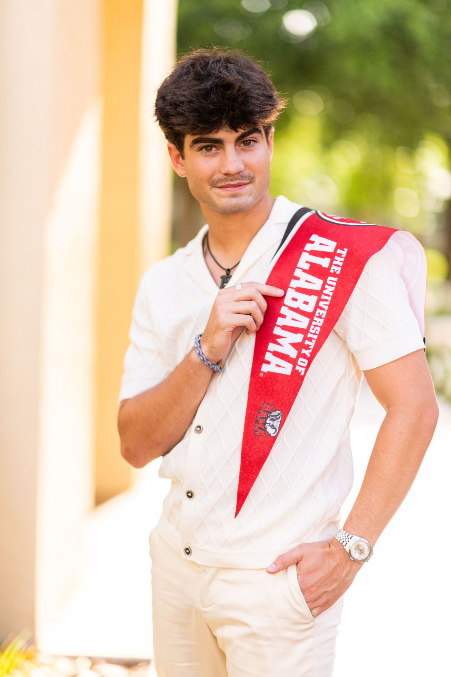 Smiling teen holding University of Alabama pennant over shoulder in white shirt outdoors.