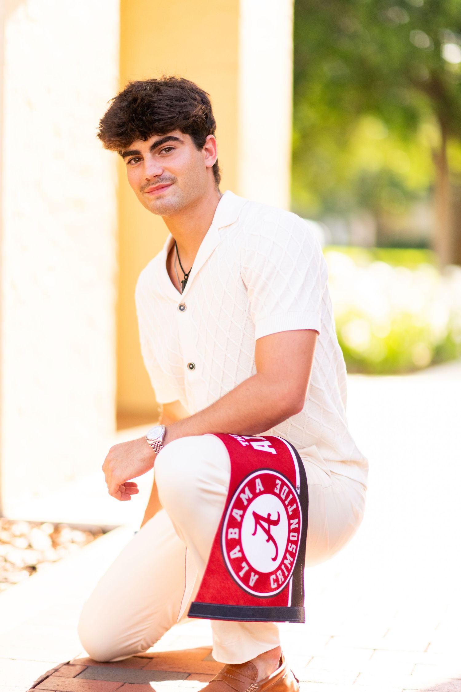 Young man in cream outfit kneeling with University of Alabama pennant in sunny outdoor setting.