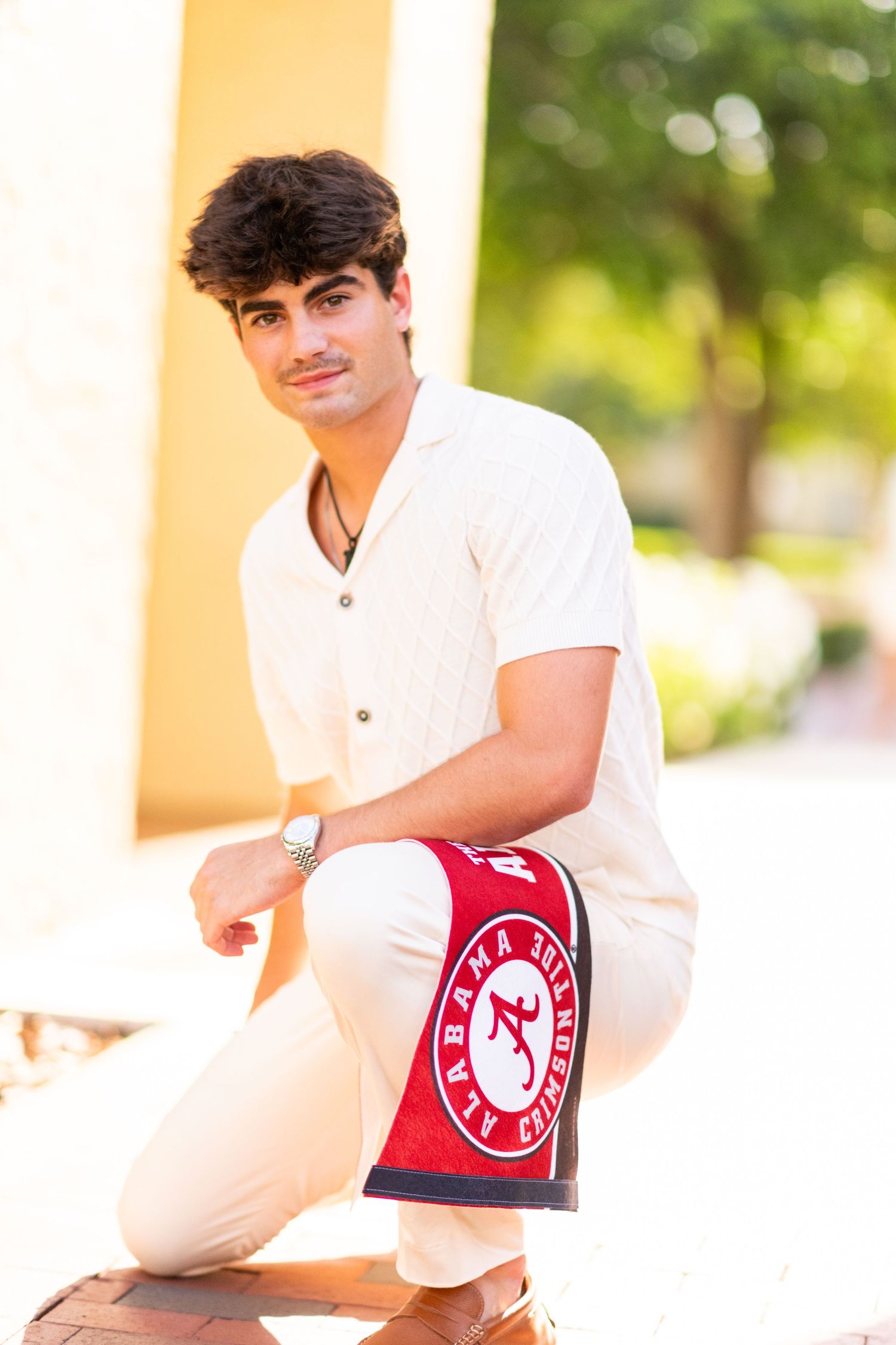 Young man in cream outfit kneeling outdoors holding a red University of Alabama pennant flag.