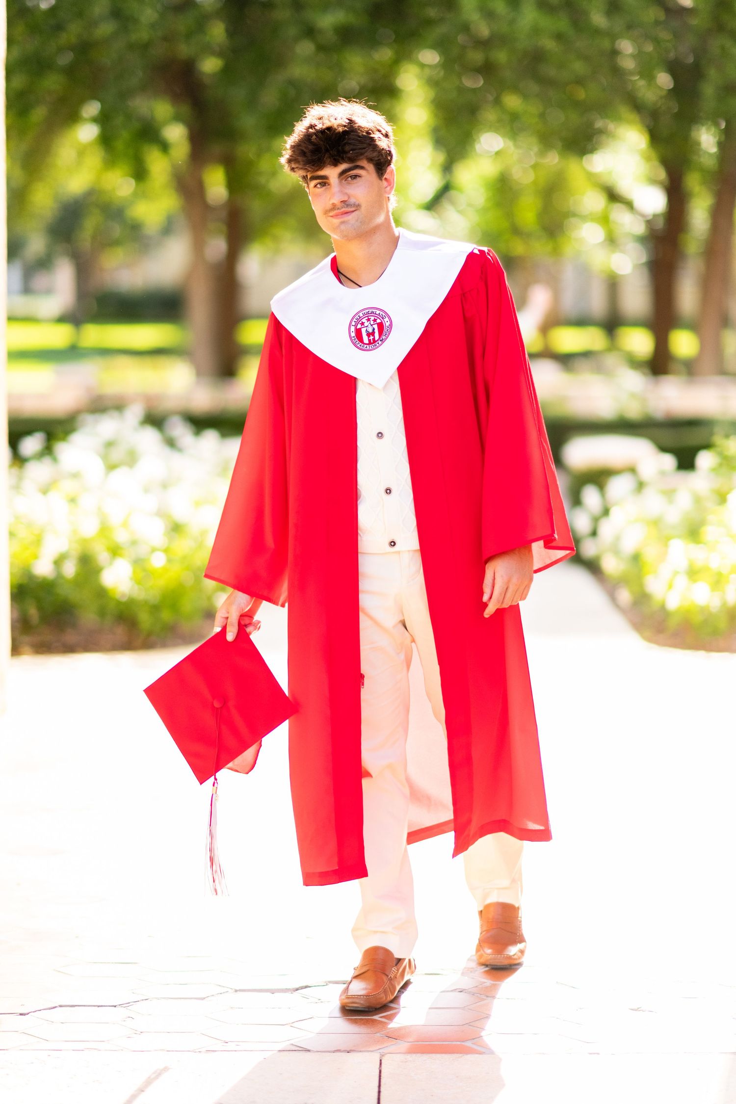 Young man walking in red graduation cap and gown holding mortarboard in tree-lined path.