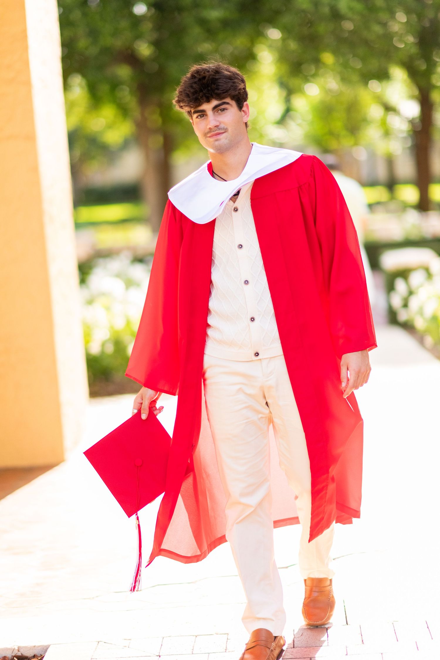 Young man in red graduation gown and white outfit walking outdoors holding cap.