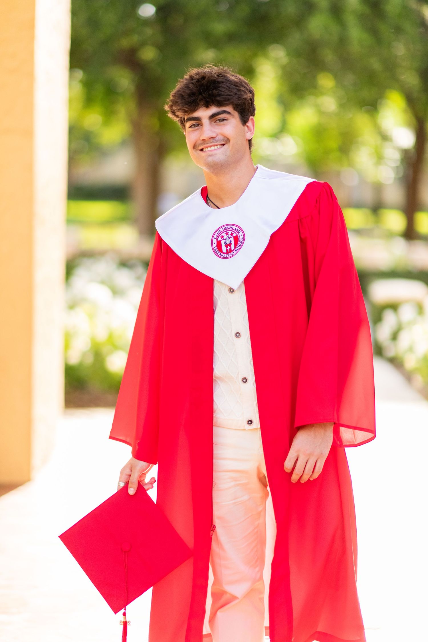 Smiling graduate in red and white cap and gown holding graduation cap outdoors.