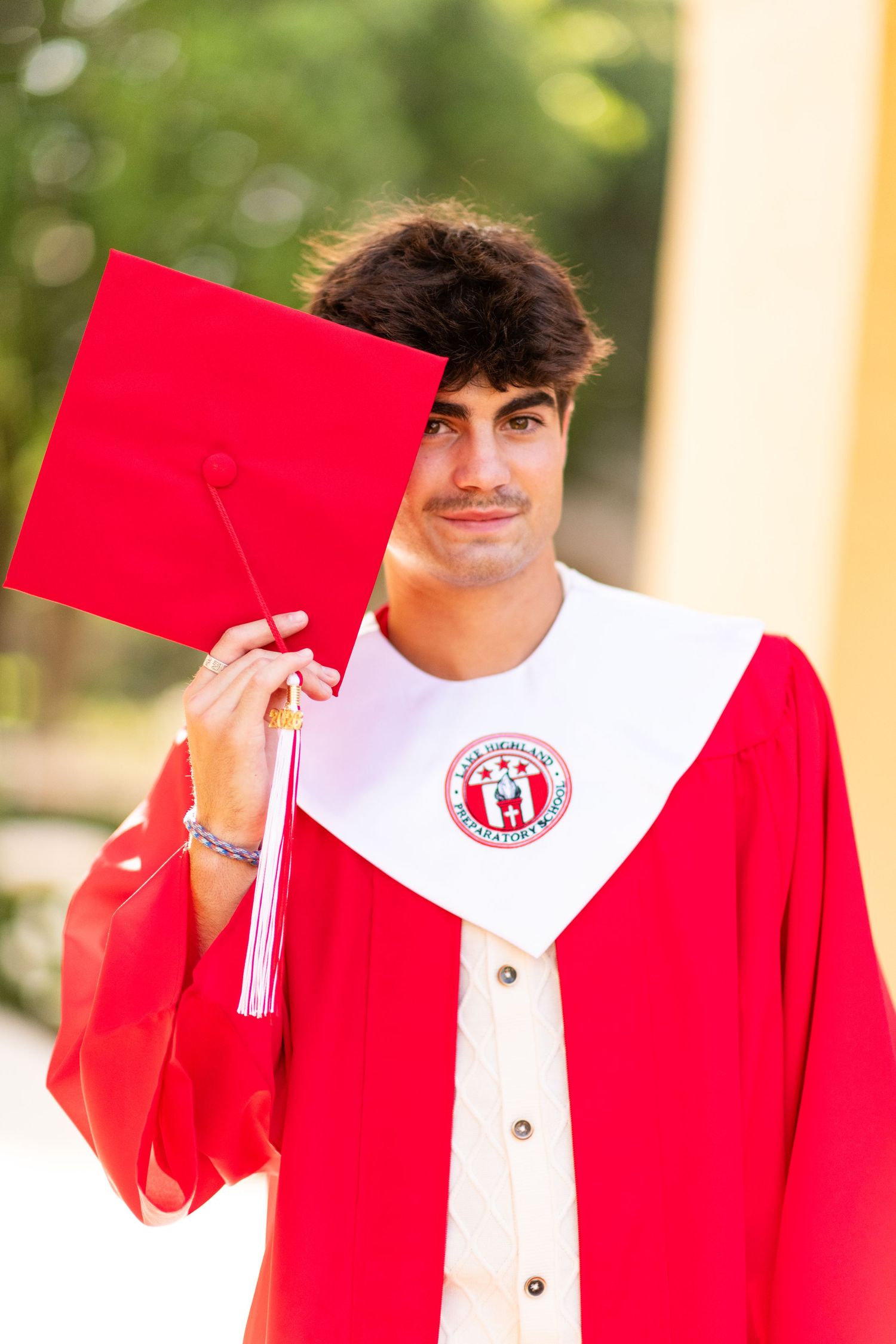 Graduating young man in red cap and gown holding mortarboard in front of his face.