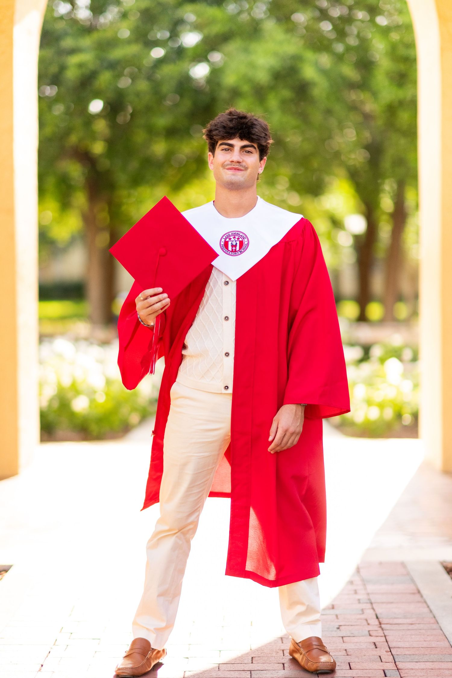 Graduating senior in red cap and gown standing under arched walkway with greenery.