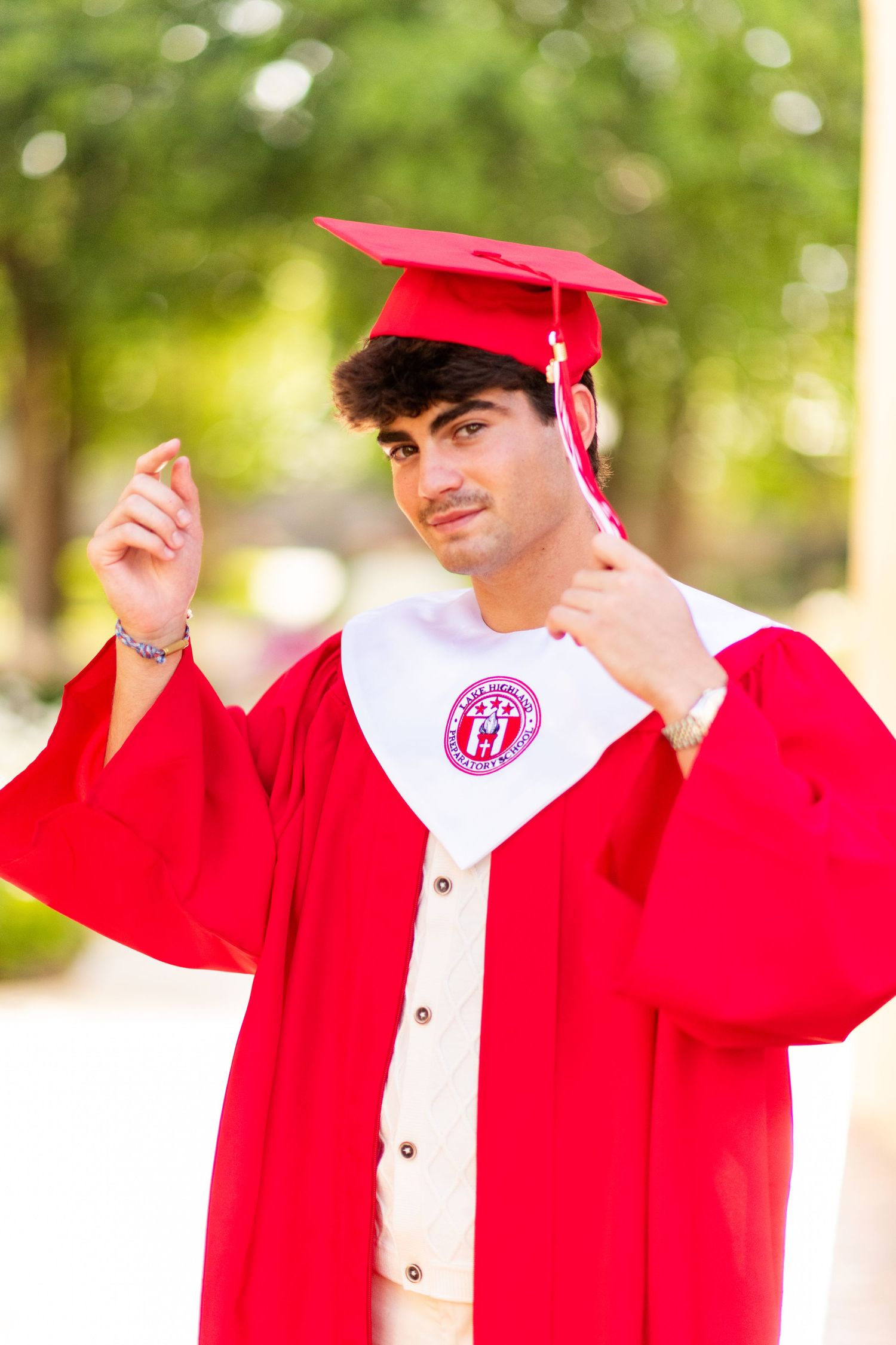 Young man in red graduation cap and gown adjusting tassel during outdoor graduation photos.