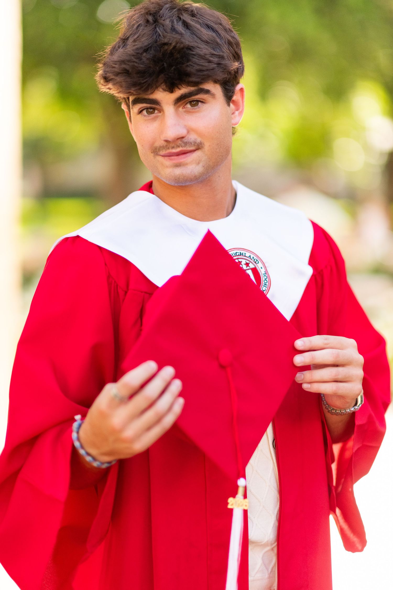 Smiling young man in red graduation gown holding cap outdoors with green trees behind him.
