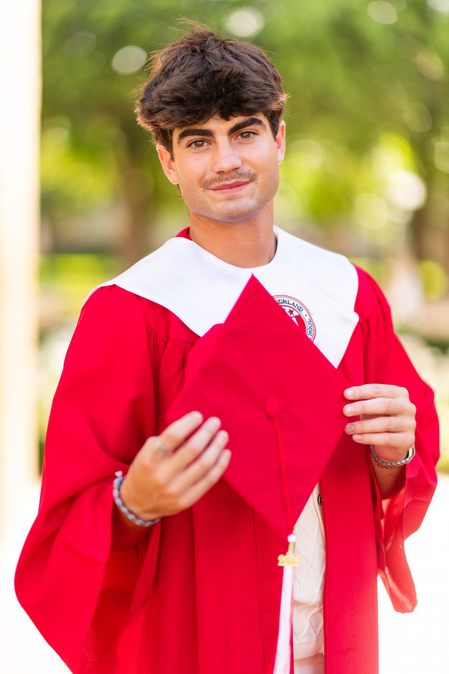 Young man in red graduation gown holding cap outdoors with green bokeh background.