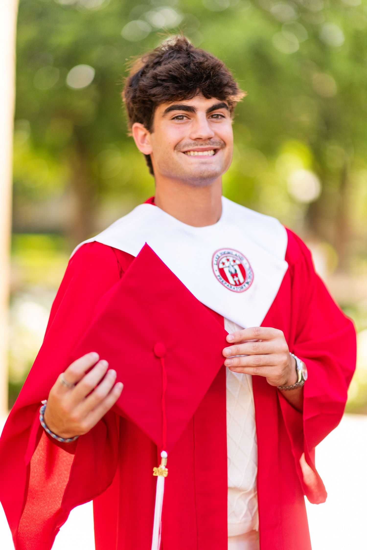 Smiling graduate in red and white cap and gown posing outdoors with green trees behind.