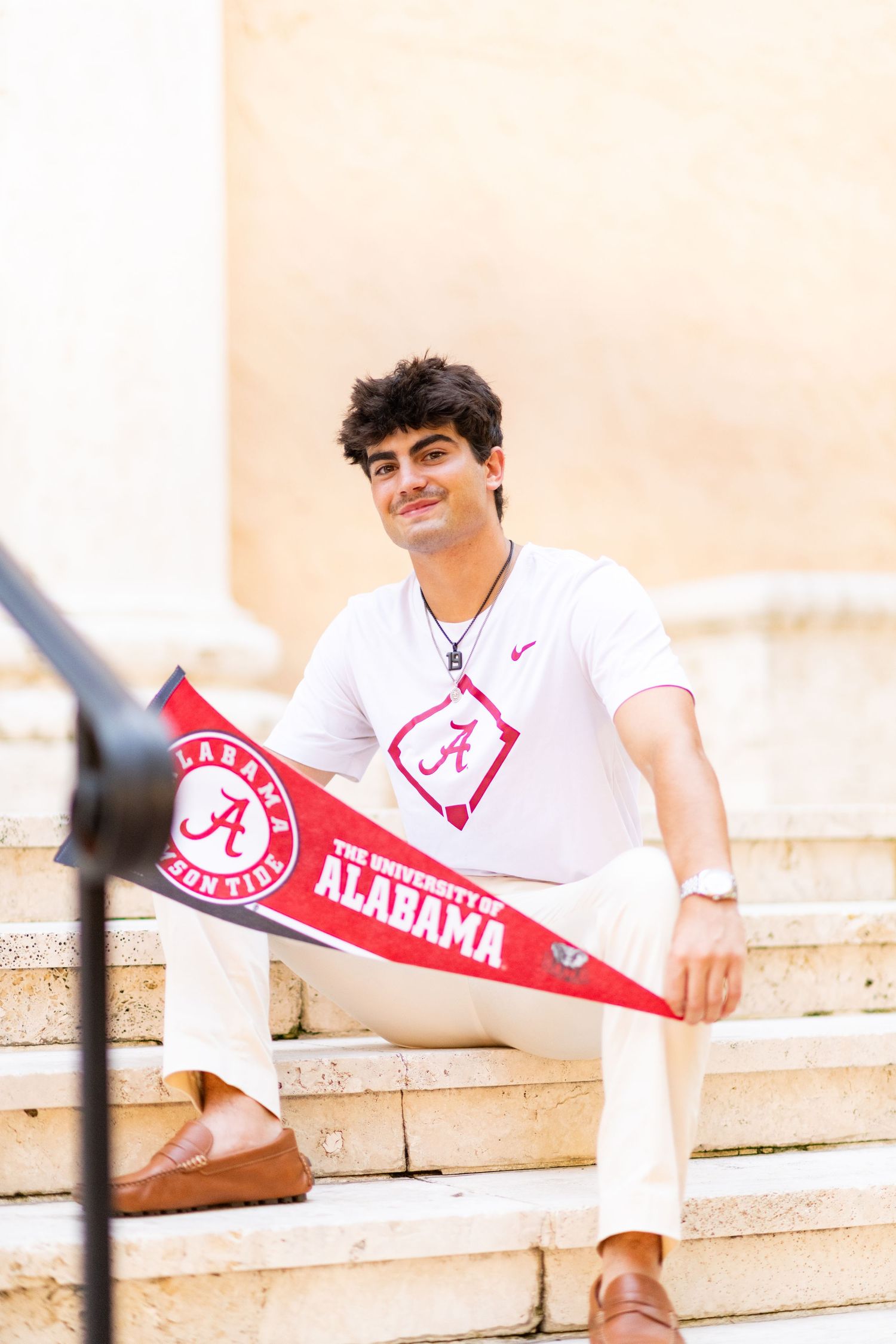 Young man in Alabama Crimson Tide shirt sitting on stone steps holding team pennants.