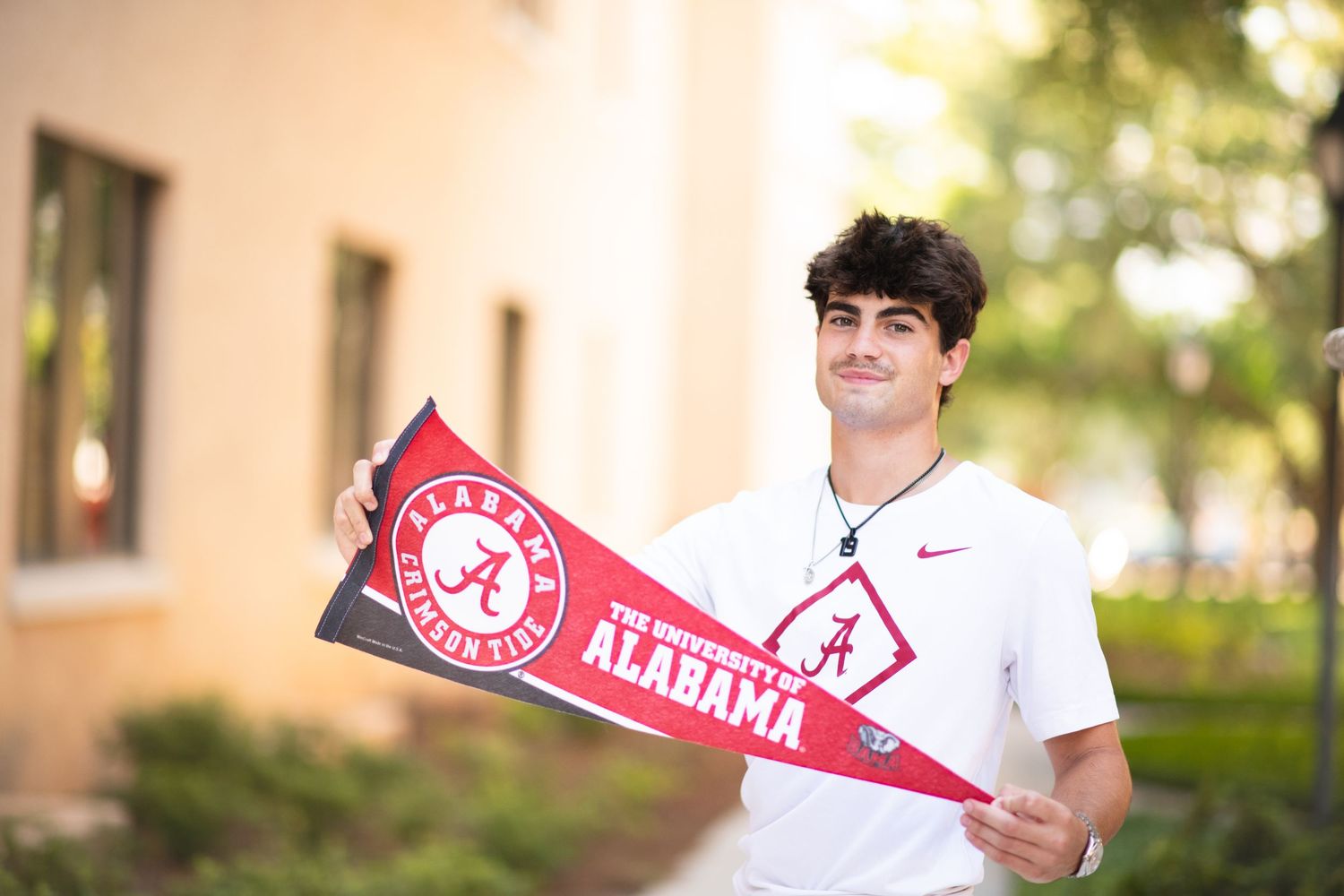 Young man holding a University of Alabama pennant flag outdoors on a sunny day wearing a white Nike shirt.
