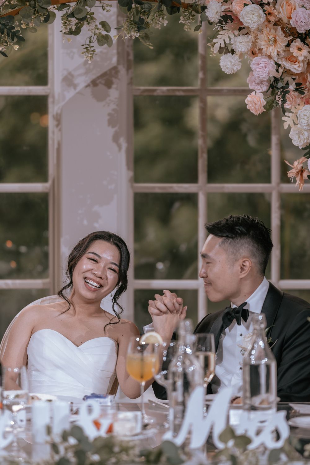 Smiling bride and groom hold hands at wedding reception table adorned with floral arrangements and champagne glasses.