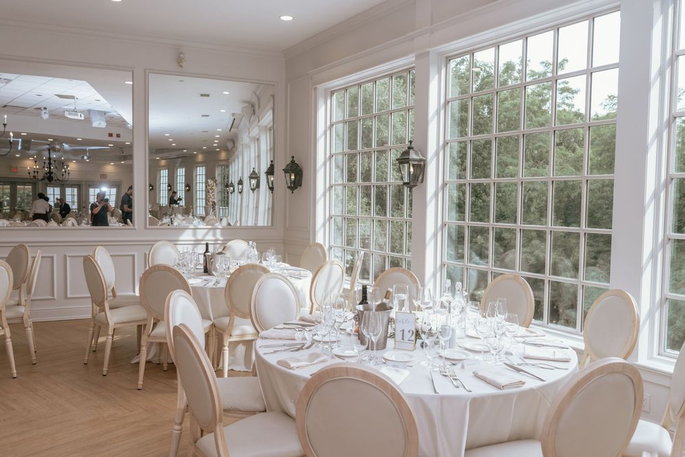 Elegant white dining room with round tables, fine glassware, cream chairs, and large windows overlooking lush greenery.