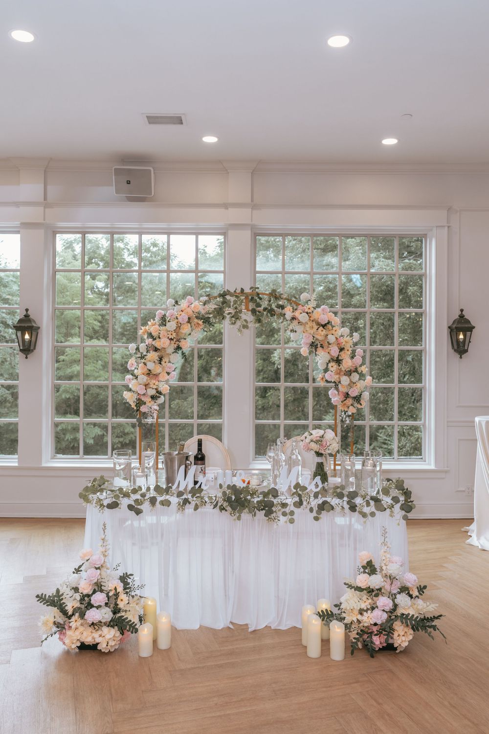 Elegant wedding sweetheart table with floral arch, candles, and blush pink roses in a bright white ballroom.