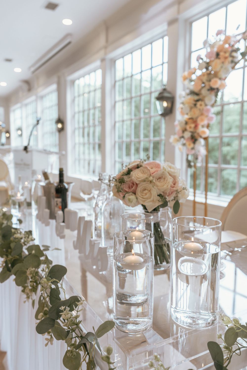Elegant wedding reception table with floating candles, white roses, and eucalyptus garland in a bright venue.