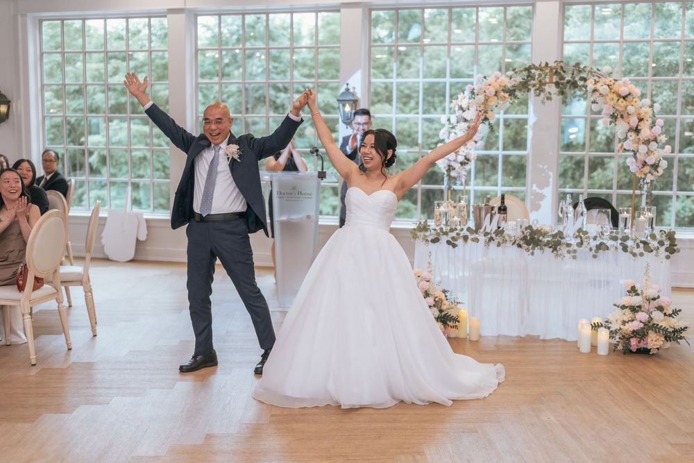 Bride and groom celebrate their grand entrance at an elegant wedding reception with floral arch and large windows.
