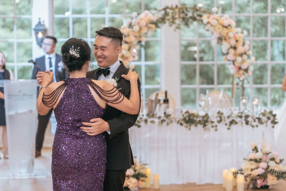 Groom in tuxedo shares a dance with his mother in purple sequin dress at a floral wedding reception.