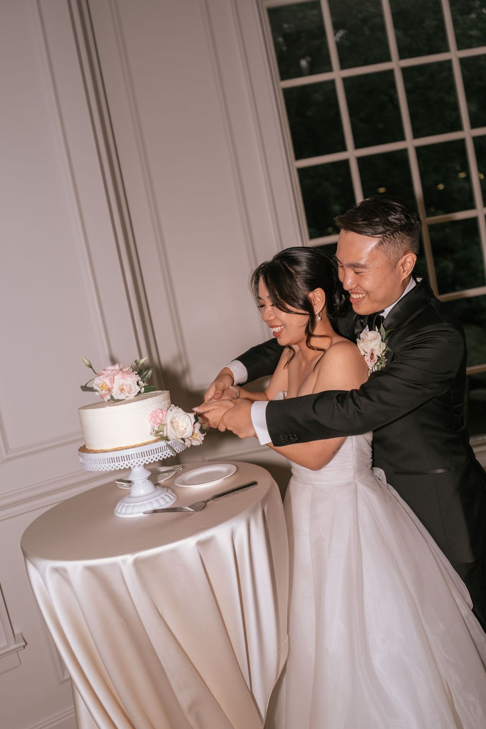 Smiling bride and groom cutting their two-tier white wedding cake adorned with pink roses at their reception.