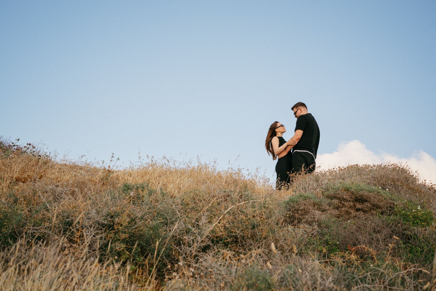Couple embracing romantically on a golden hillside under a clear blue sky in an outdoor engagement photo.