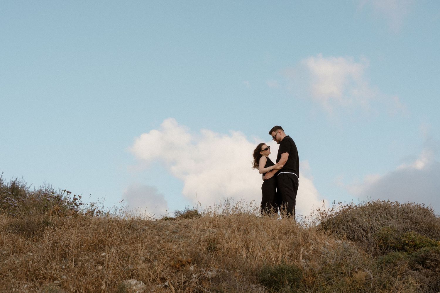 Couple embracing romantically on a golden hillside under a clear blue sky in an outdoor engagement photo.
