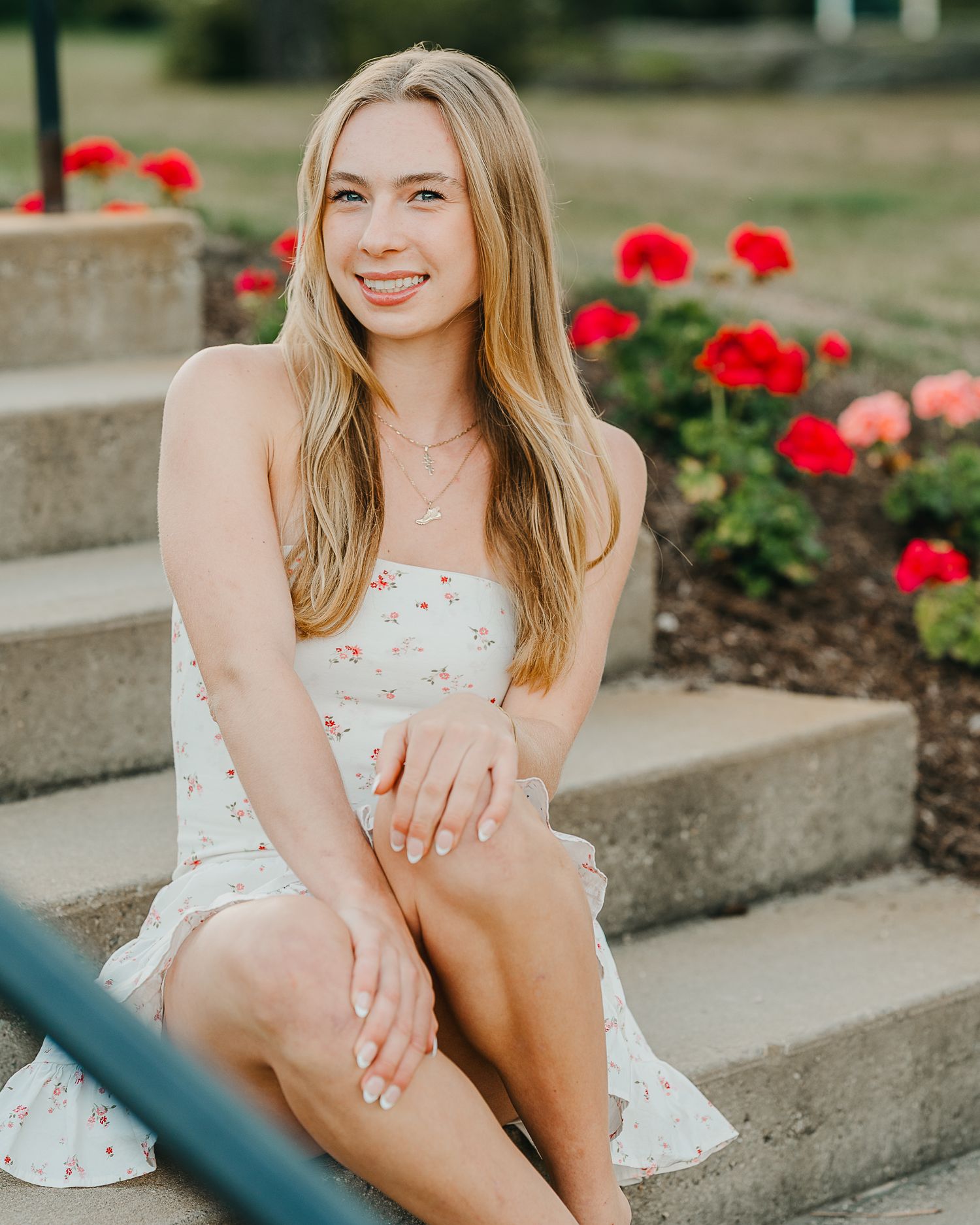 high school senior in a short fun white dress sitting barefoot on concrete pillar with flowers behind her