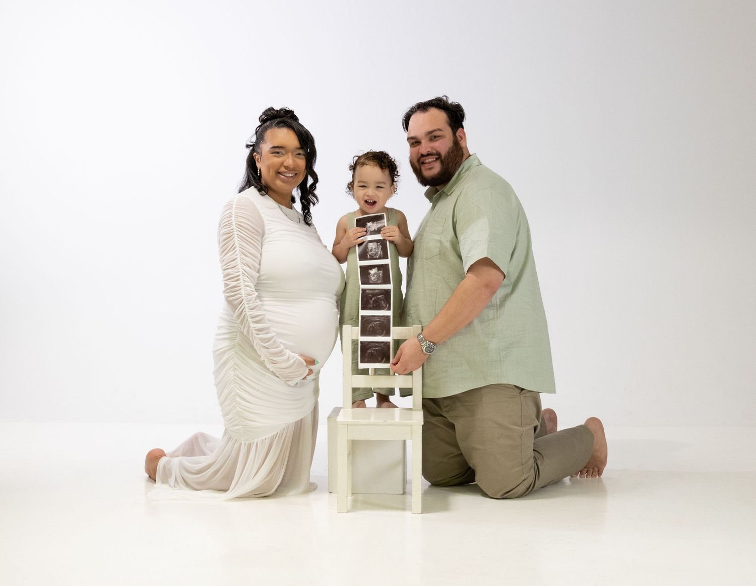Pregnant woman in white lace dress poses with toddler and husband holding ultrasound photos on white chair.