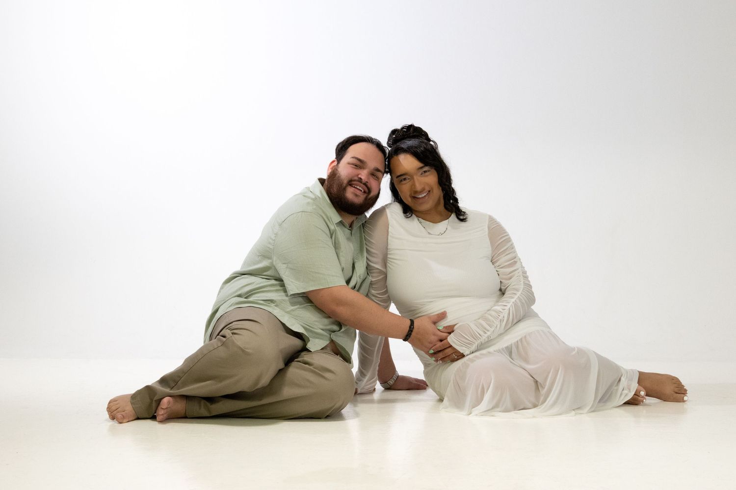 Smiling couple sitting on white floor, pregnant woman in white dress, man in green shirt, maternity photo session.
