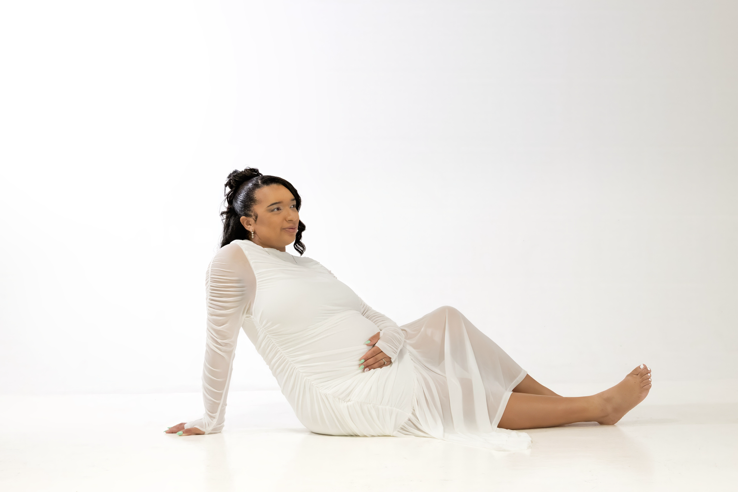 Pregnant woman in white sheer dress sitting gracefully on white studio floor, barefoot with braided hair.