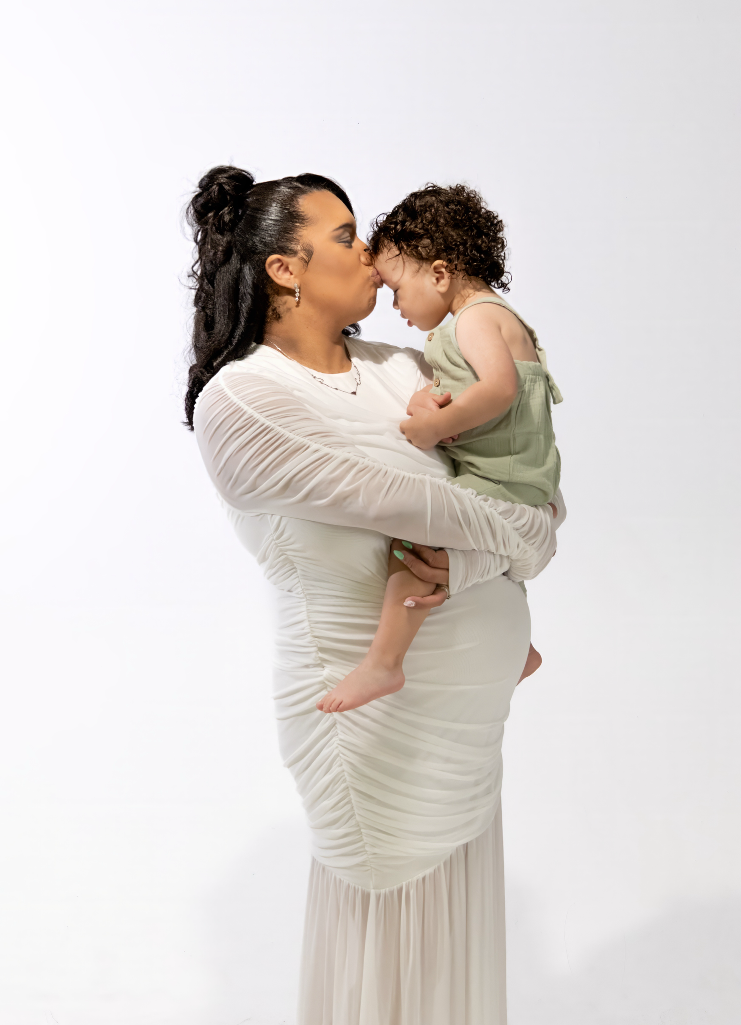 A mother in a white dress kisses her curly-haired baby in a green outfit against a white background.