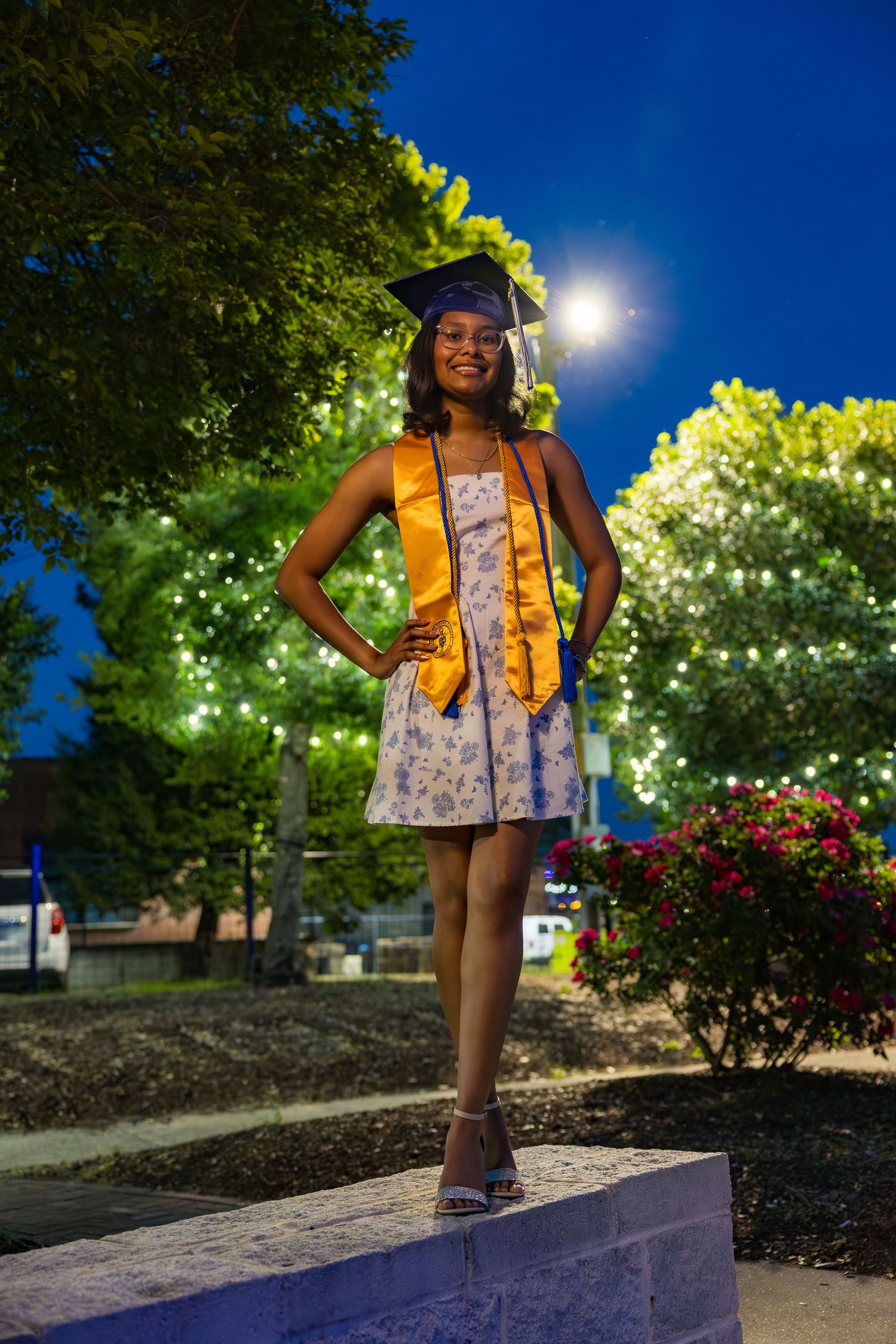 Young woman in graduation cap and gold stole poses outdoors at night with festive lights in background.