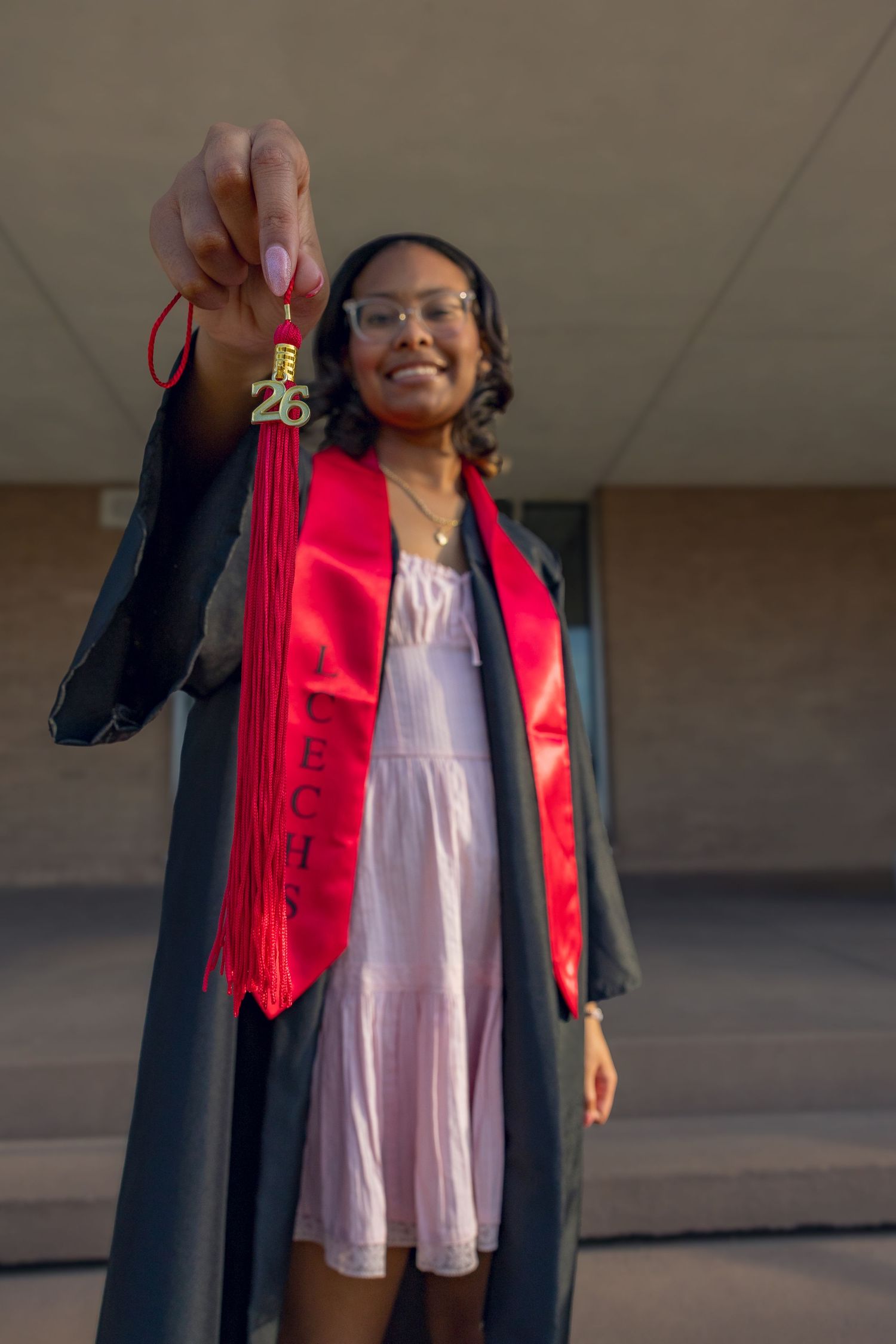 Smiling graduate in black gown and red sash holds up tassel, celebrating class of 2026.