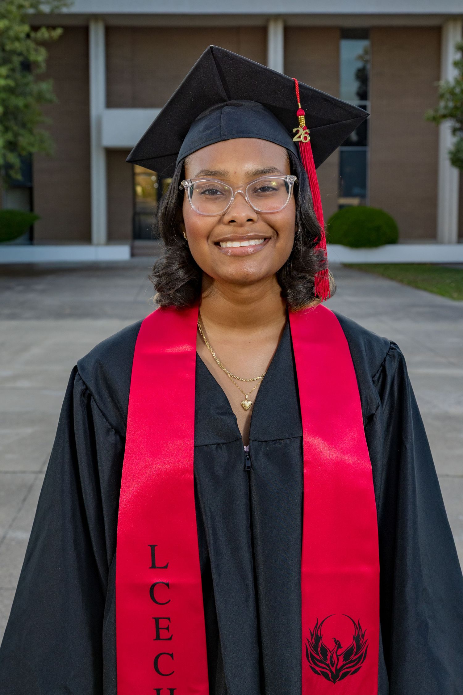 Smiling graduate in black cap and gown with red LCEC honor stole poses outdoors at graduation ceremony.