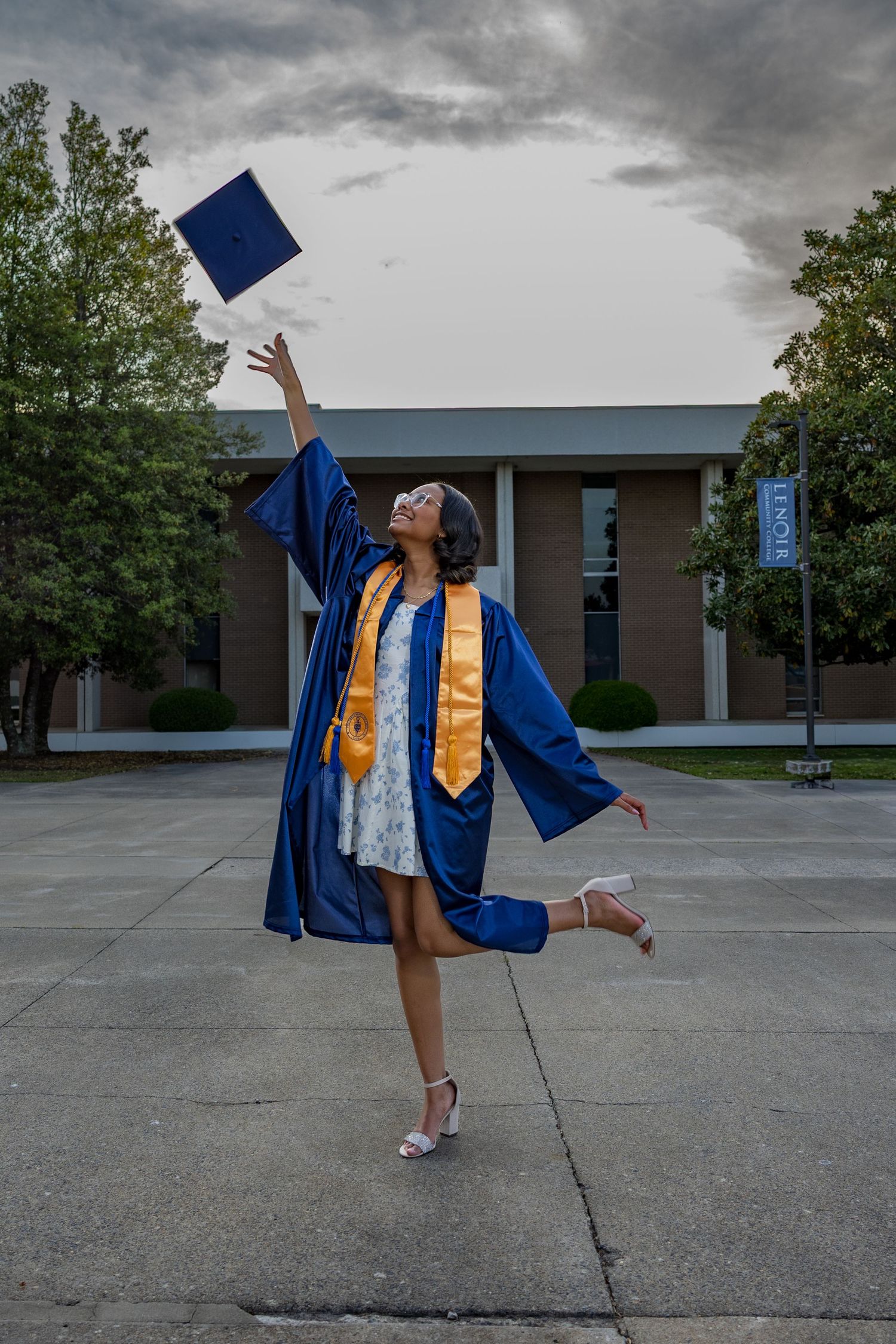 A joyful graduate in blue cap and gown tosses her mortarboard into the air while posing on campus.